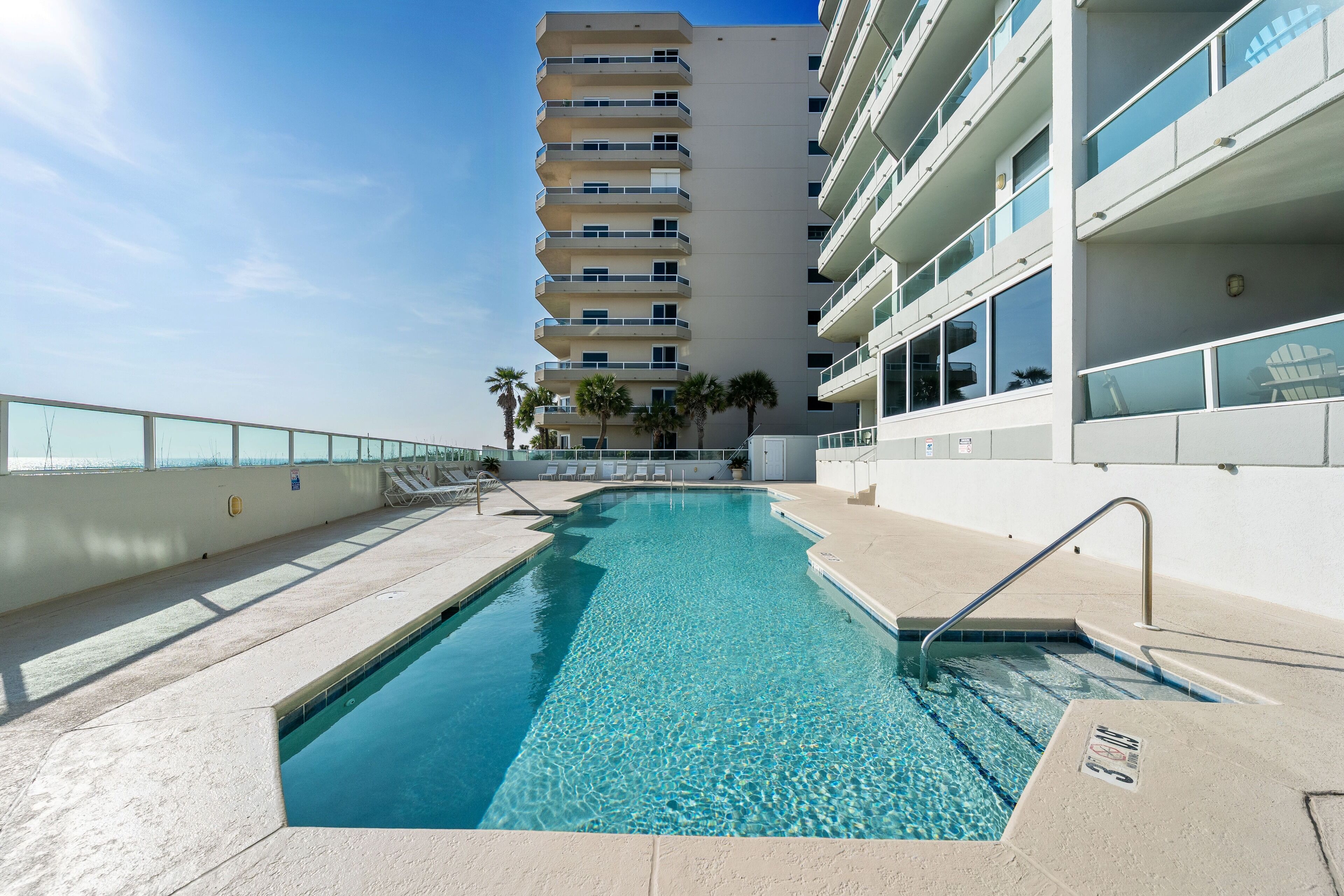A swimming pool with a clear blue water, surrounded by a poolside area with lounge chairs and a building with multiple balconies in the background.