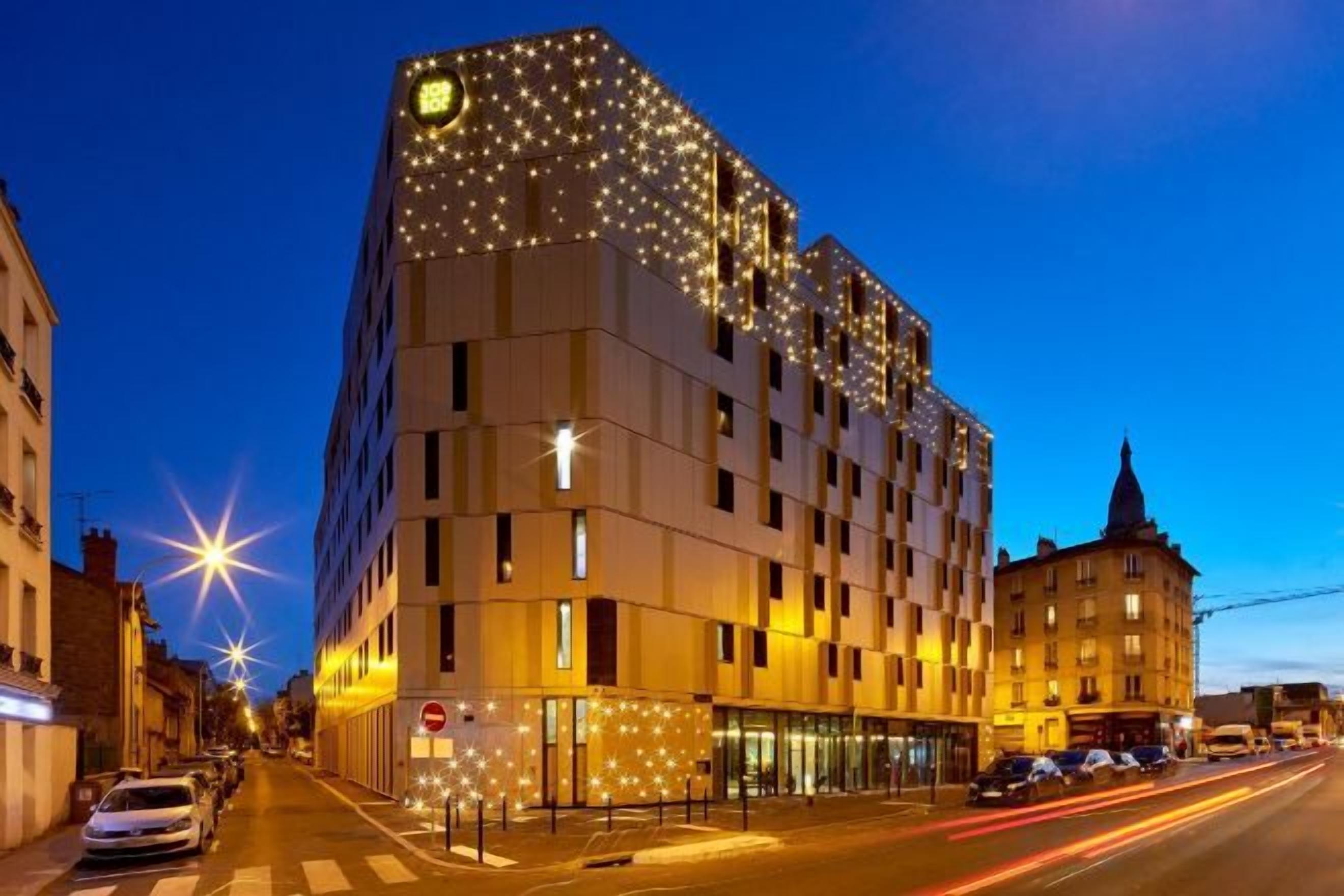 Un hôtel à plusieurs étages, doté d’un design moderne, avec un bâtiment d’angle remarquable, des fenêtres illuminées et une entrée bien visible.