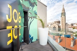 Un balcon donnant sur un paysage urbain, avec notamment une vue sur le clocher d’une église, et un mur orné d’un grand logo.
