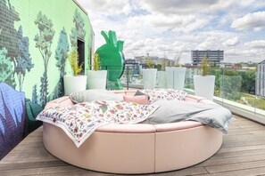 Un espacio de descanso circular y rosado con una manta estampada, una escultura abstracta verde y vistas a la ciudad.