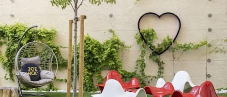 Un jardín con una silla colgante, sillas rojas y blancas, y una decoración con forma de corazón en una pared.