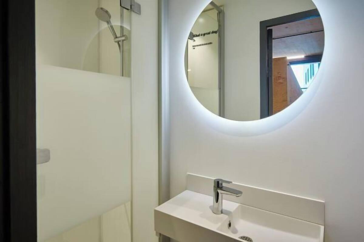 Un baño moderno con un lavamanos blanco, un espejo redondo y una ducha.
