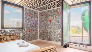 Une pièce avec un lit, une fenêtre et un mur orné d’un motif labyrinthique et de motifs en forme de cœur.