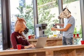 Una persona con máscara de caballo usando una computadora portátil en una barra de madera con una máquina de café y tazas rojas.