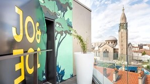 Un balcon donnant sur un paysage urbain, une grande plante en pot et un immeuble surmonté d’une tour.