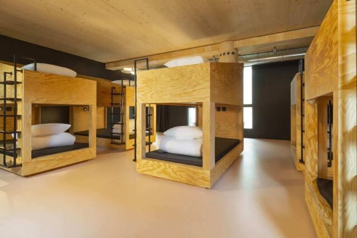 Camas literas con estructuras de madera y colchones negros, dispuestas en fila sobre un suelo de madera y un techo oscuro.