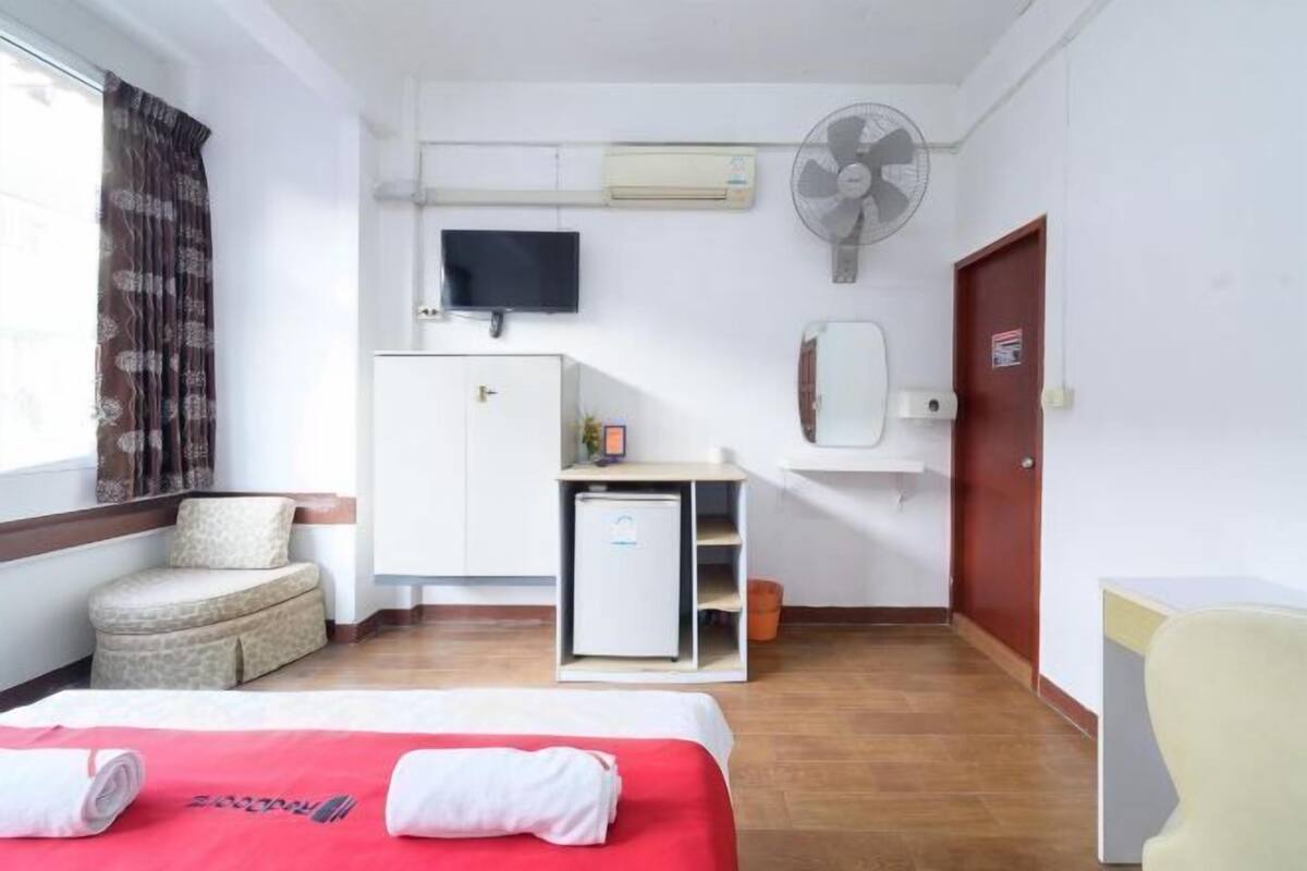 A hotel room with a bed, a desk, a small refrigerator, a TV, a fan, a mirror, and a window with curtains.