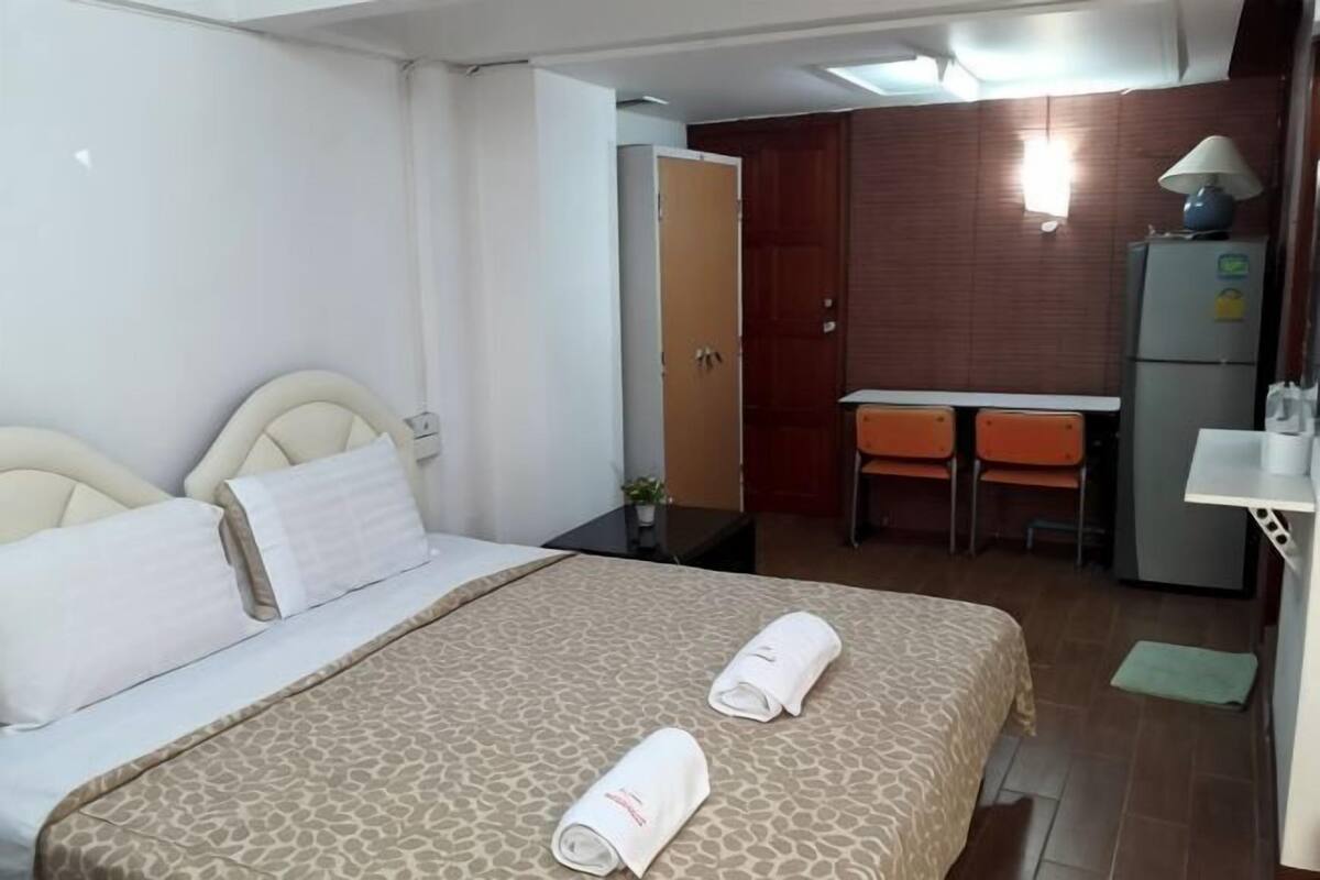 A hotel room with a bed, a desk, a chair, a refrigerator, and a small table with a lamp.