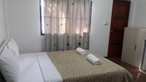 A neatly made bed with a patterned bedspread, a window with curtains, and a wooden door. - Noy's House (Bangkok)