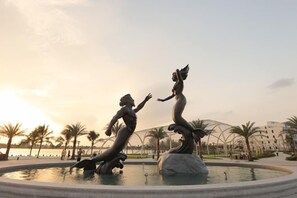 A fountain with two statues of figures in flowing poses, surrounded by palm trees and a body of water, with buildings in the background. - Sunshine Apartment - Vinhomes Ocean Park Ha Noi (Hanoi)
