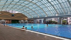 An indoor swimming pool with a glass ceiling, surrounded by lounge areas and a wooden deck. - Sunshine Apartment - Vinhomes Ocean Park Ha Noi (Hanoi)