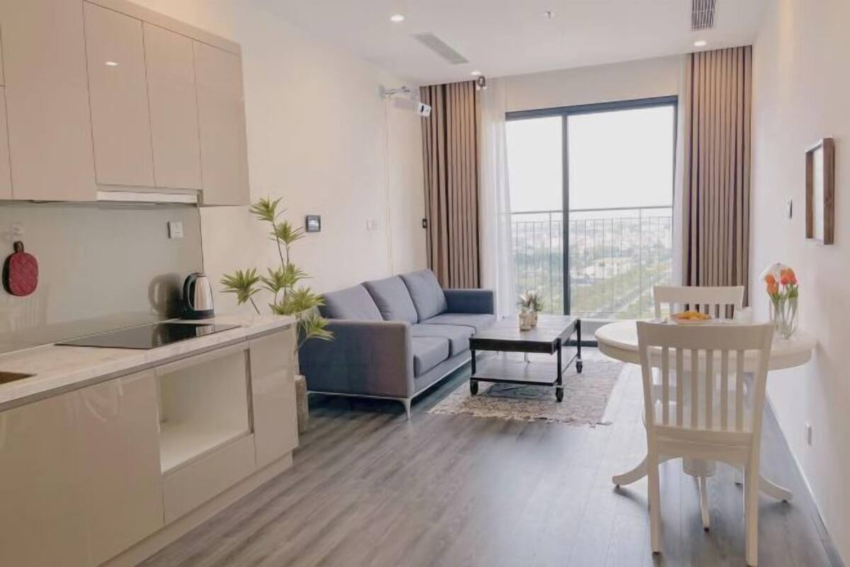 A modern apartment with a kitchen, a grey sofa, a dining table with a white chair, and a large window with curtains.