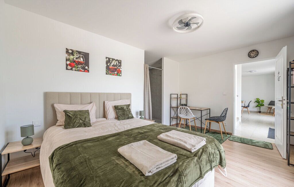 A bedroom with a bed, two pillows, a green blanket, a bedside table with a lamp, and a chair.