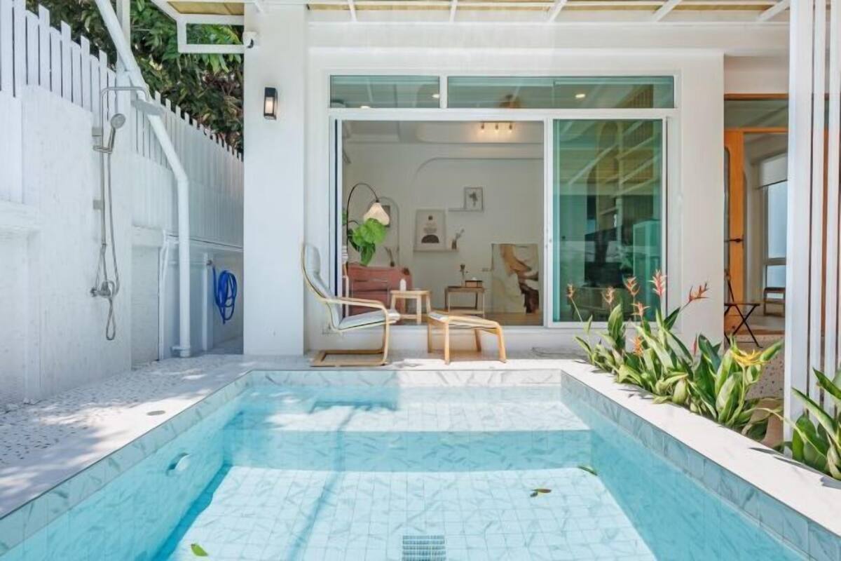 Een zwembad met betegeld terras, een wit hekwerk en een glazen deur die leidt naar een kamer met een stoel en een tafel.