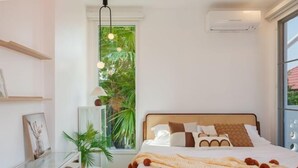 Une chambre moderne avec un grand lit, un bureau en verre, une plante en pot et une vue sur la verdure par la fenêtre.