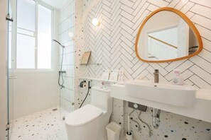 Une salle de bain avec un sol carrelé de carreaux blancs, des toilettes blanches, une douche en verre, un lavabo blanc avec un miroir et un mur orné d’un motif en chevrons.