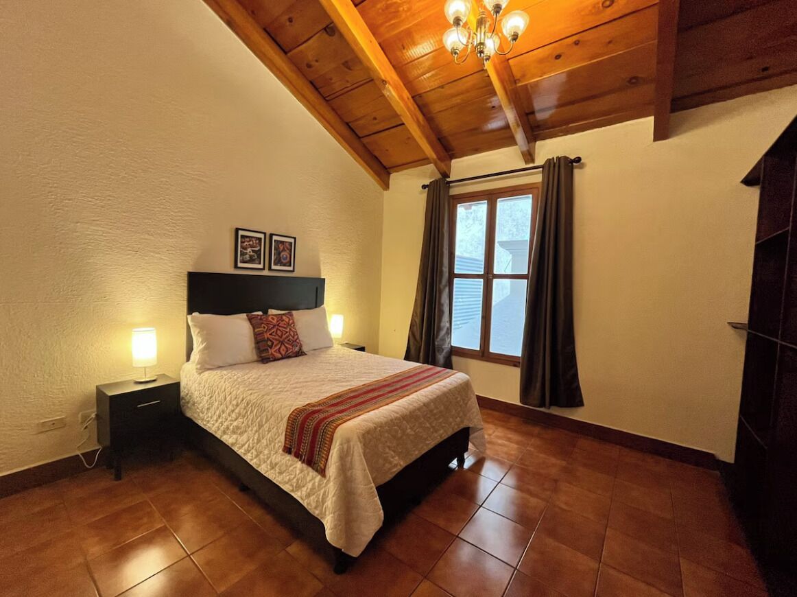 A bedroom with a wooden ceiling, a bed with a white bedspread, a nightstand with a lamp, and a window with curtains.