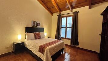 A bedroom with a wooden ceiling, a bed with a white bedspread, a nightstand with a lamp, and a window with curtains.