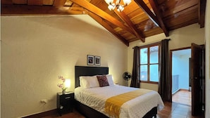 A bedroom with a wooden ceiling, a bed with a white duvet and a yellow throw, a nightstand with a lamp, and a window with curtains. - Excellent location in Antigua for 4 guests (Antigua Guatemala)