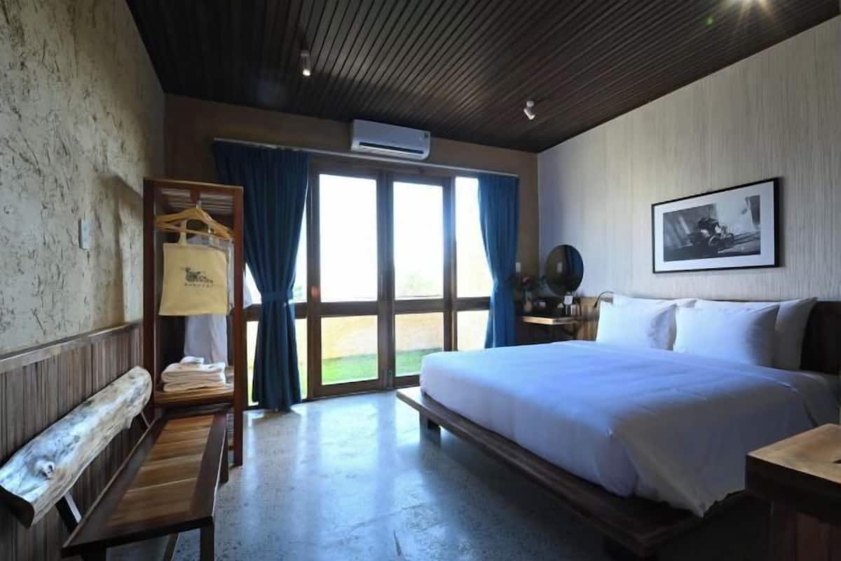 A bedroom with a large bed, a wooden bench, a wardrobe, and a window with curtains.