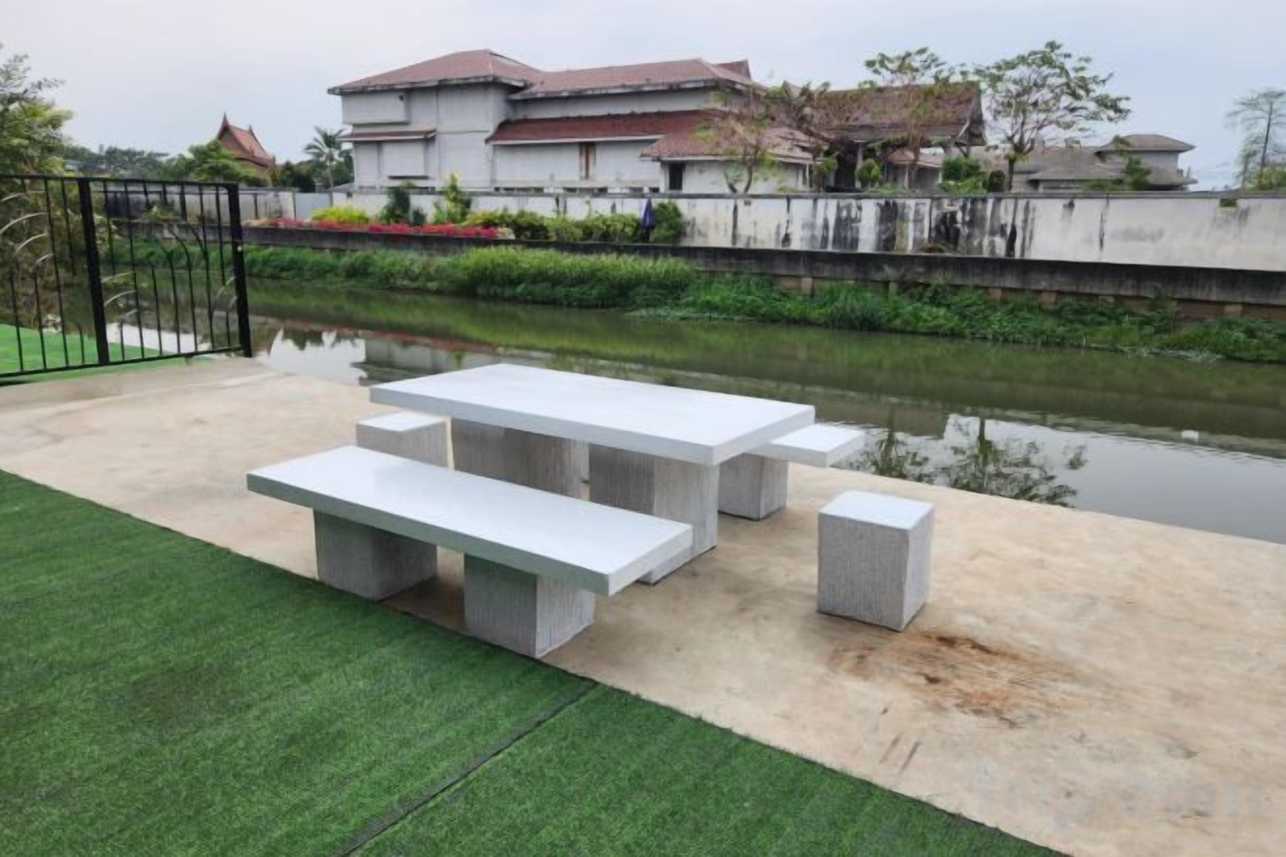 Un patio exterior con una mesa y bancos de concreto, un área con césped y un cuerpo de agua al fondo.