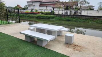 Un patio exterior con una mesa y bancos de concreto, un área con césped y un cuerpo de agua al fondo.