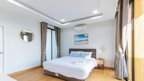 A hotel room with a bed, a nightstand, a wall-mounted air conditioner, a painting, and a window with curtains. - Riverfront Pool Villa Rangsit Klong 4 (Thanyaburi)