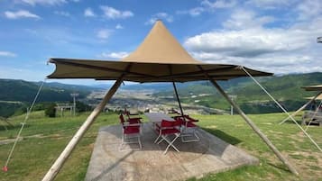 A camping setup with a conical tent, picnic table, and chairs on a grassy hill with a view of the valley.