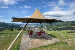 Un campamento con una tienda cónica, mesa de picnic y sillas sobre una colina cubierta de pasto, con vista al valle.