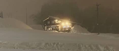 A snow-covered landscape with a building featuring a porch and illuminated windows.
