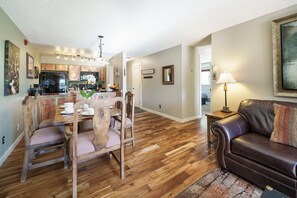 Un coin repas avec une table et des chaises en bois, une cuisine à l’arrière-plan, un salon avec un canapé en cuir et un couloir menant à une autre pièce.