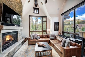 A spacious living room with a stone fireplace, leather sofas, and large windows offering a mountain view.