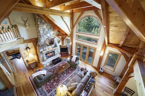 A spacious living room with a stone fireplace, wooden beams, and large windows offering views of the outdoors.