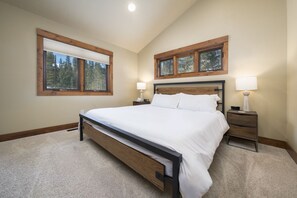 A bedroom with a large bed, two bedside tables with lamps, a window with blinds, and a view of trees.
