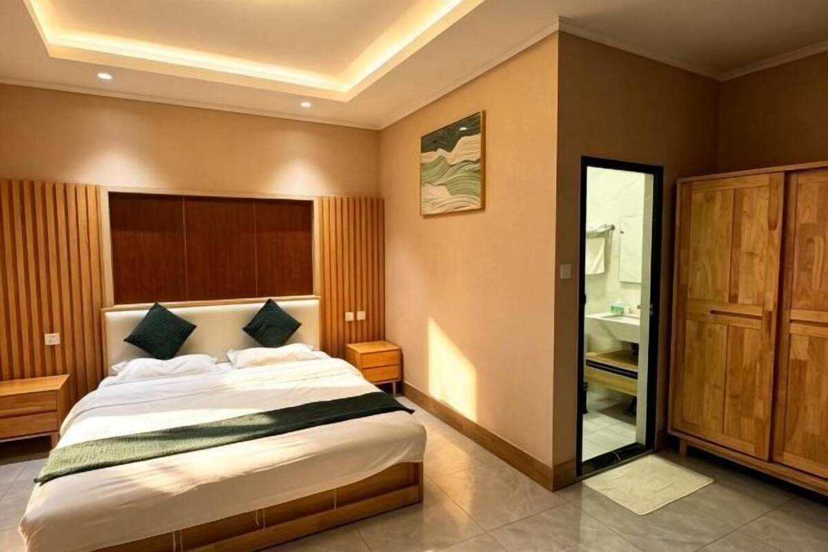 A bedroom with a bed, wooden headboard, bedside tables, a wardrobe, and a bathroom visible through a glass door.
