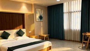 Ein Hotelzimmer mit Bett, Nachttisch, Sessel, einem Fenster mit Vorhängen und einem Wandbild.