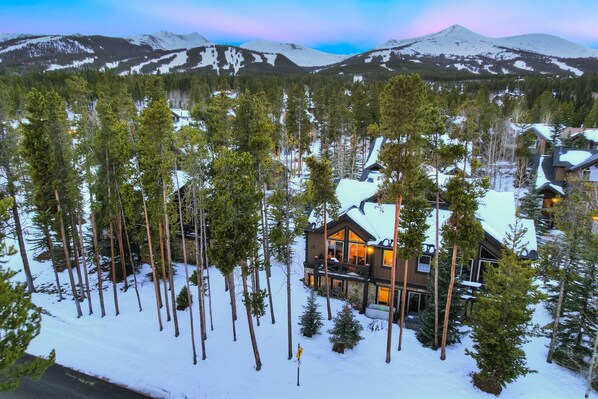 Un chalet enneigé, avec une façade en pierre et de nombreuses fenêtres, entouré de grands pins.