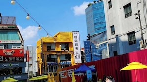 A street scene with a yellow building, a blue glass building, and a red building. There are yellow umbrellas and people walking. - CareCove (Seoul)