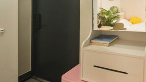 A bedroom with a bed, bedside table, and a small shelf with a plant and books. - CareCove (Seoul)