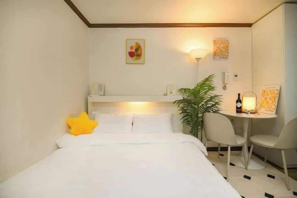 A hotel room with a bed, a desk, chairs, a plant, and wall art.
