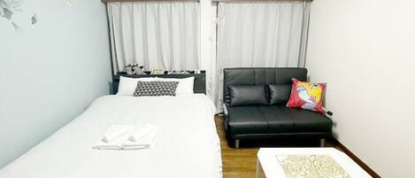 A hotel room with a bed, a black leather sofa, a small white table, and a window with curtains.