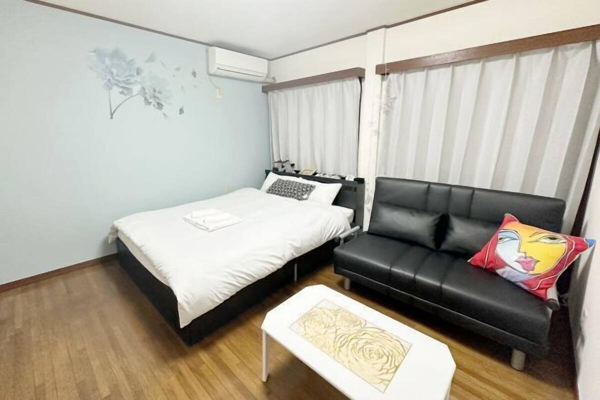 A bedroom with a bed, a black leather sofa, a small white table, and a wall-mounted air conditioner.