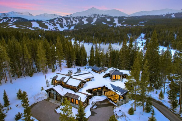 A snow-covered lodge with multiple cabins, surrounded by dense forest and mountains in the background.