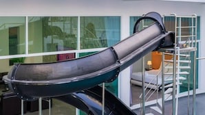 Een modern interieur met een opvallende zwarte en groene glijbaanconstructie, een glazen trap en uitzicht buiten door grote ramen.