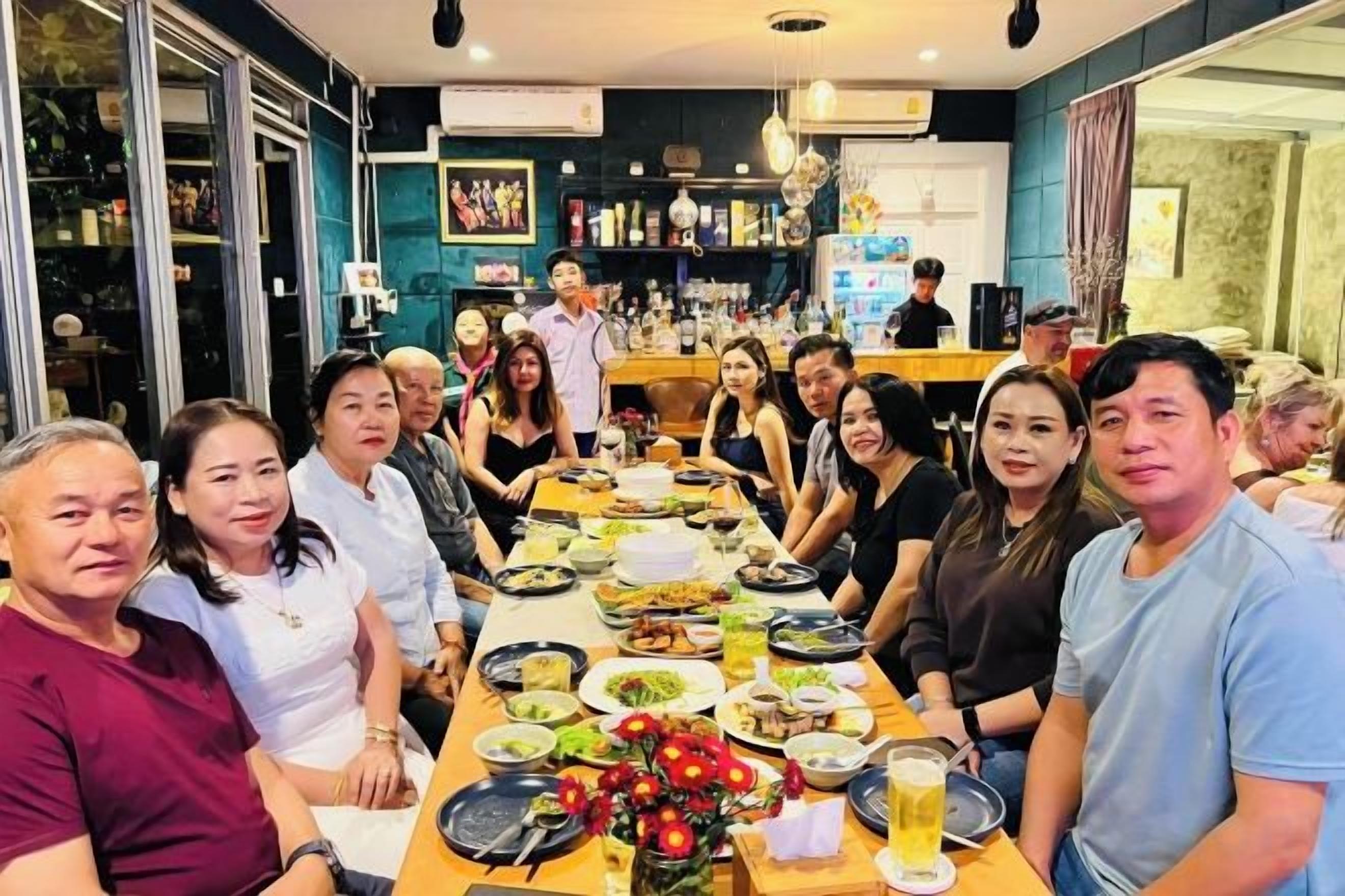 Un grupo de personas sentadas alrededor de una mesa con diversos platos y bebidas, en una habitación con una zona de bar al fondo.