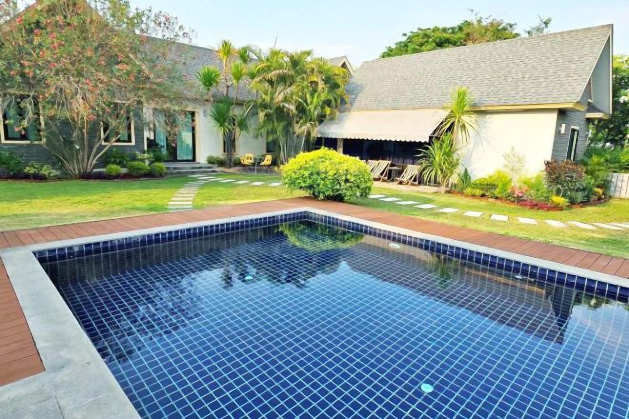 A swimming pool with a tiled edge, surrounded by a grassy area and a house with a sloped roof.