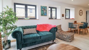 A living room with a green sofa, a red blanket, and a leopard print pouf. There is a dining area with a table and chairs, and a large window with a view. - Ewe Barn (St Austell)