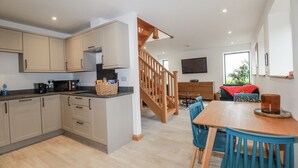 A modern kitchen with wooden cabinets, a dining area with blue chairs, and a staircase leading to a living room. - Ewe Barn (St Austell)