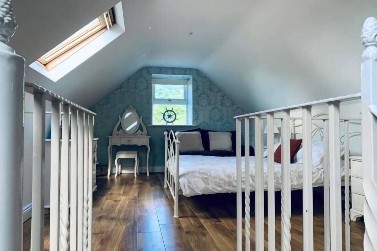 A bedroom with a sloped ceiling, a bed, a desk, and a window with a view.