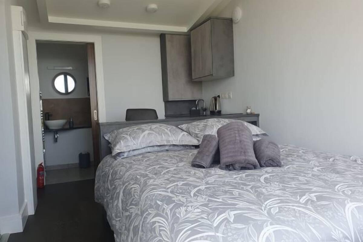 A bedroom with a bed, a small desk, a chair, a round mirror, and a built-in sink.